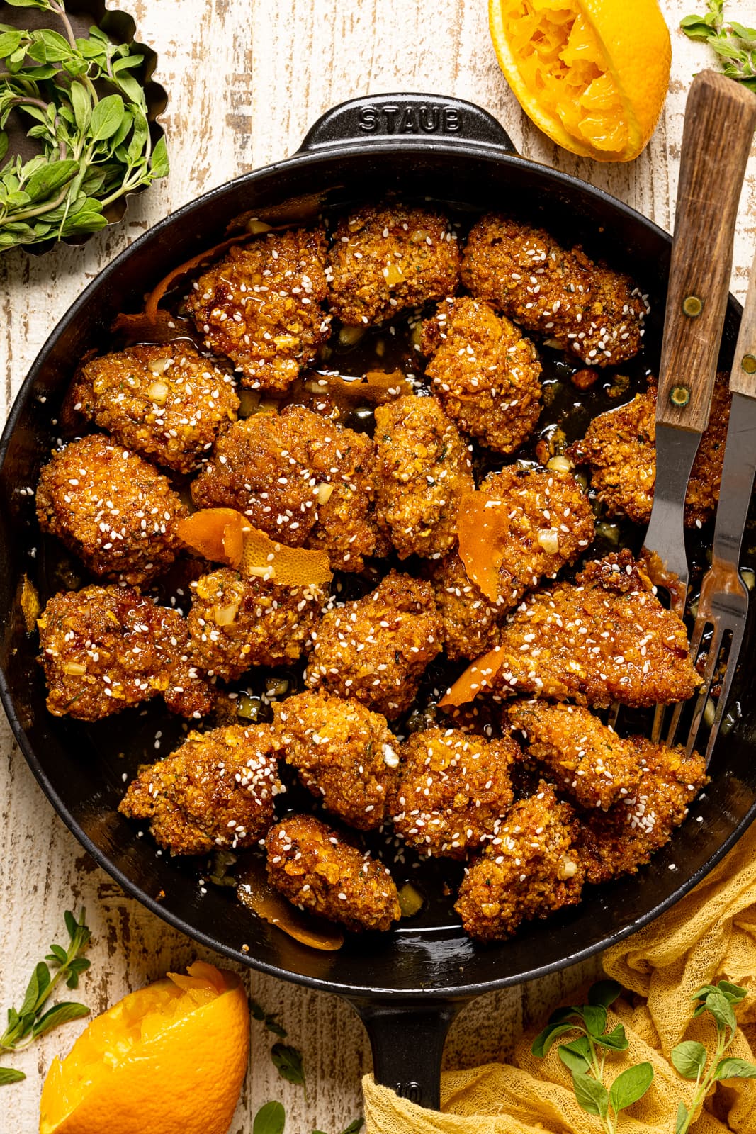 Popcorn chicken in a black skillet on a white wood table with oranges and herbs. 