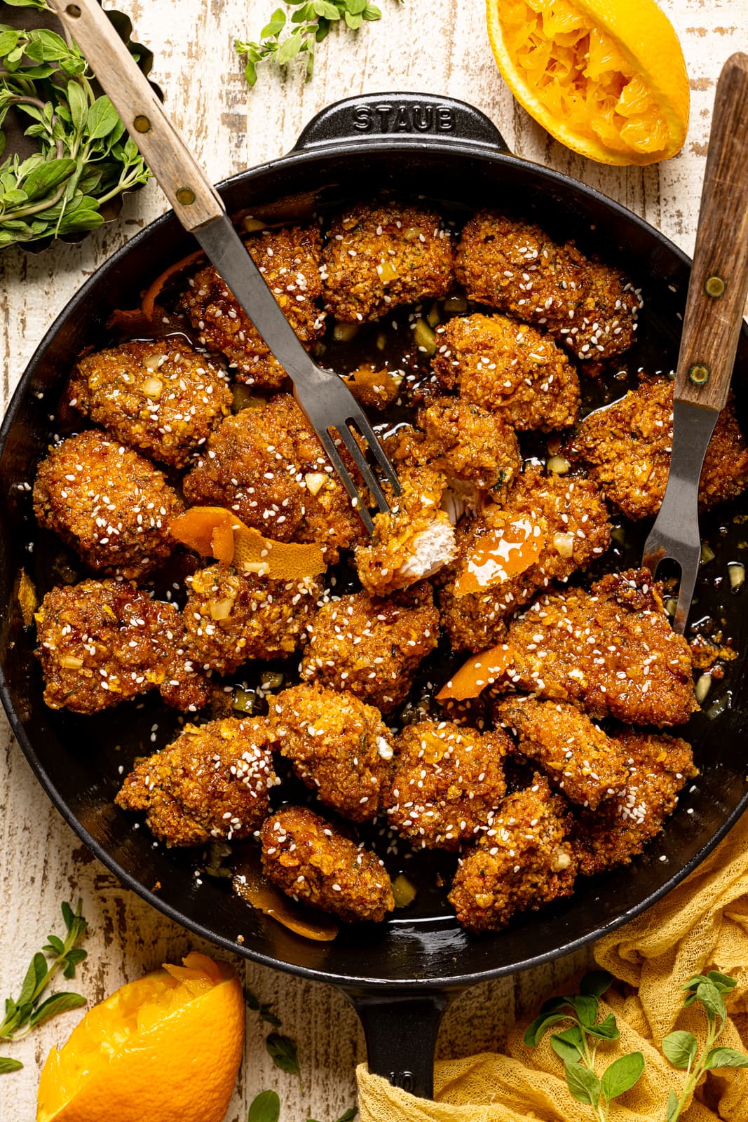 Chicken in a black skillet with two forks on a white table with oranges and herbs.