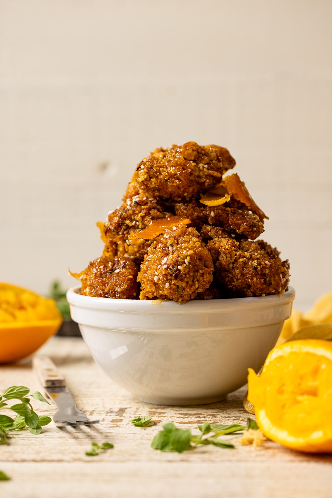 Chicken stacked in a white bowl on a white wood table with oranges and herbs. 