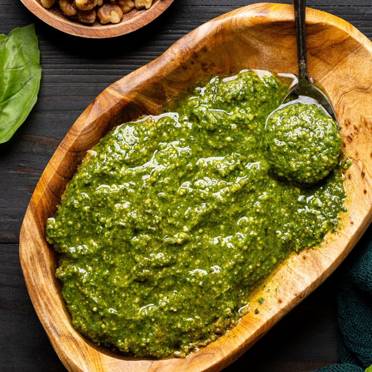 Pesto sauce in a brown wood bowl with a spoon on a black wood table with walnuts and basil leaves.