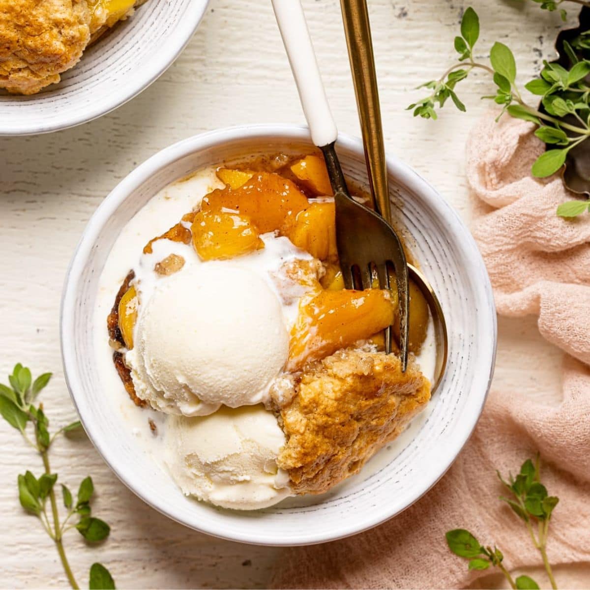 Peach cobbler in a white bowl on a white wood table next to a plate and baking dish and herbs.