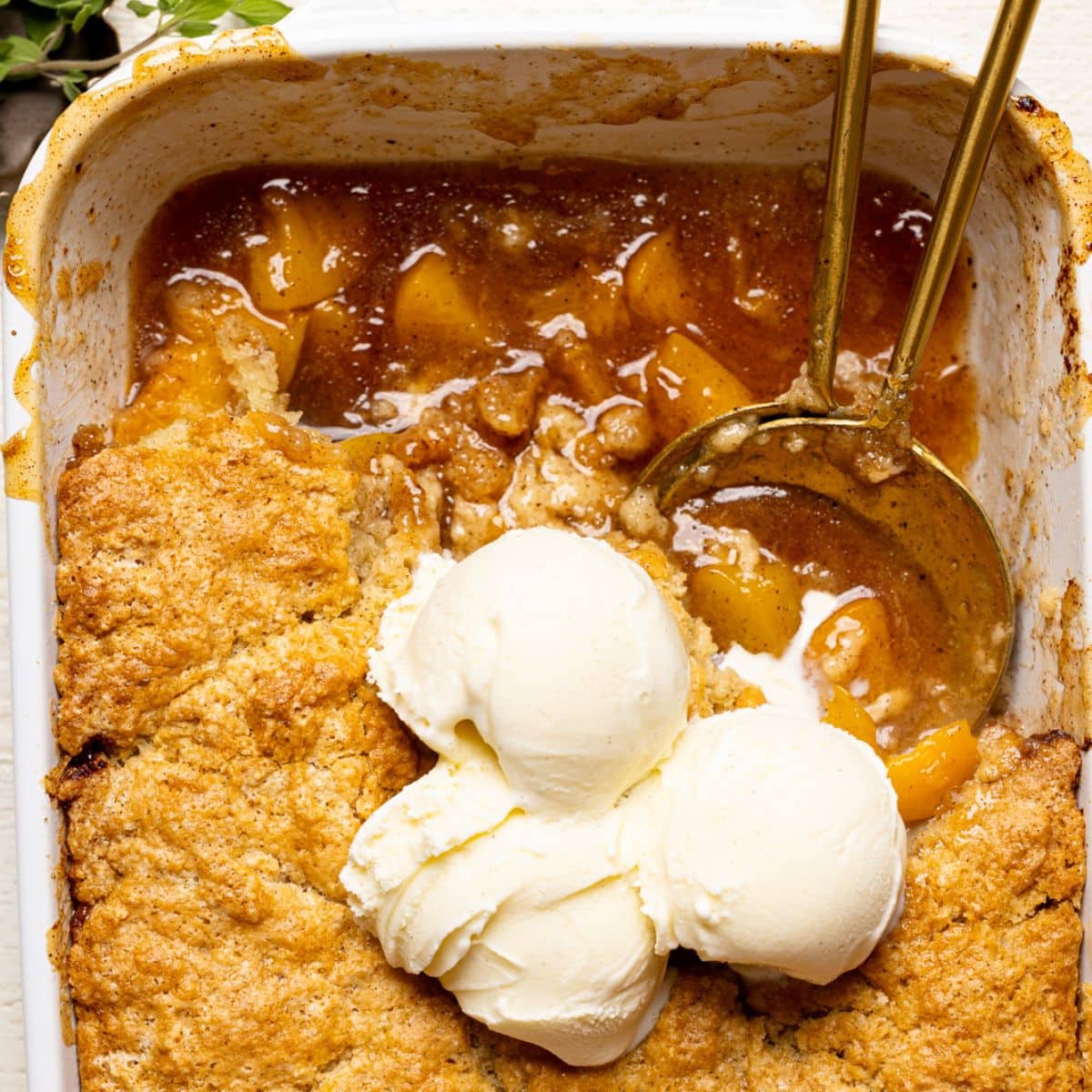 Up close shot of peach cobbler in baking dish with scoops of ice cream on top and two serving spoons.