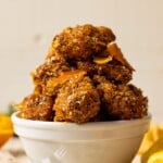 Up close shot of chicken stacked in a white bowl on a white wood table with oranges and herbs.