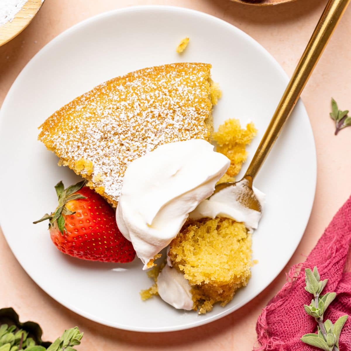 Up close shot of slice of cake on a white plate with whipped cream, strawberries, and fork on a peach colored table.