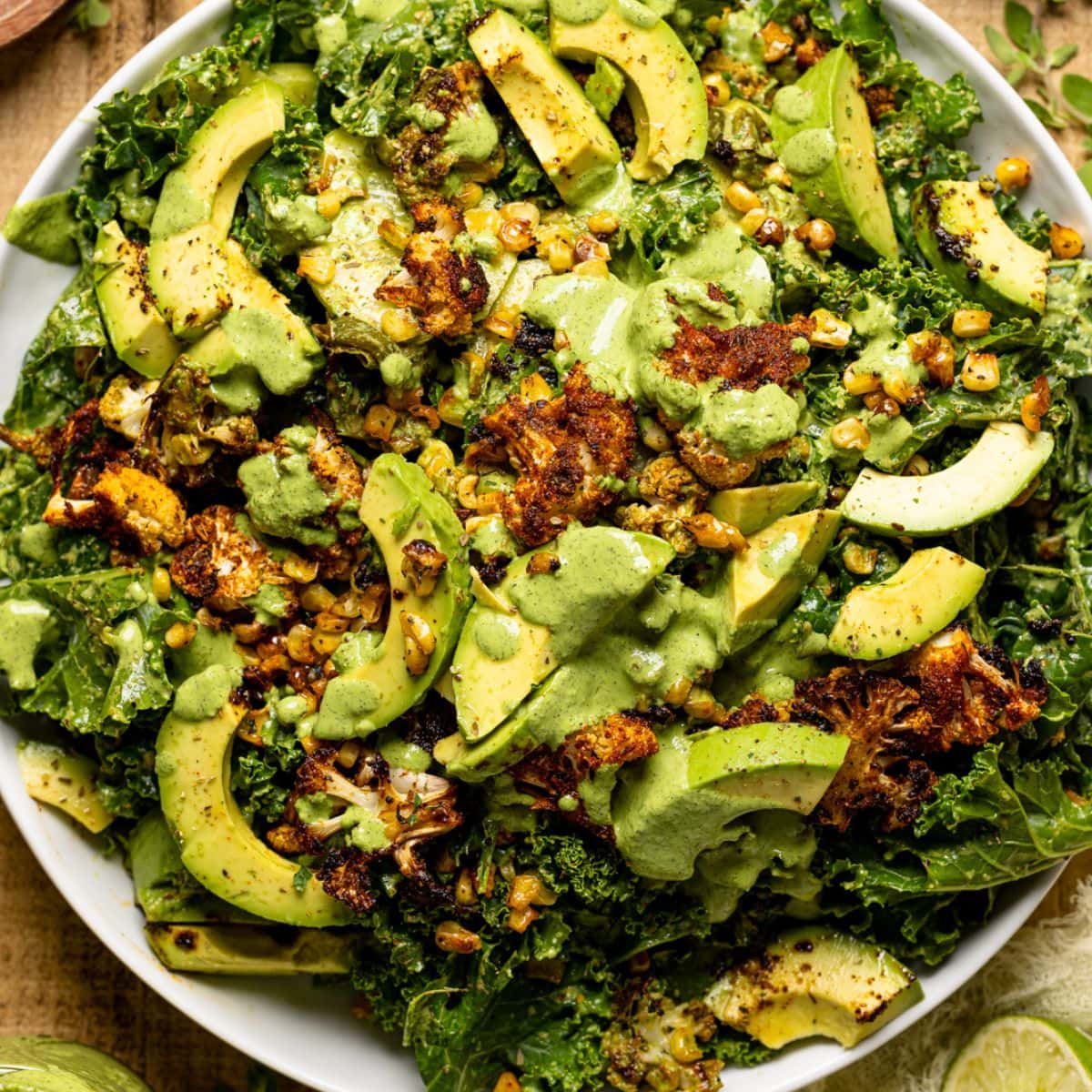 Salad on a white round plate with seasonings + herbs, lime wedges, dressing, and yellow napkin.