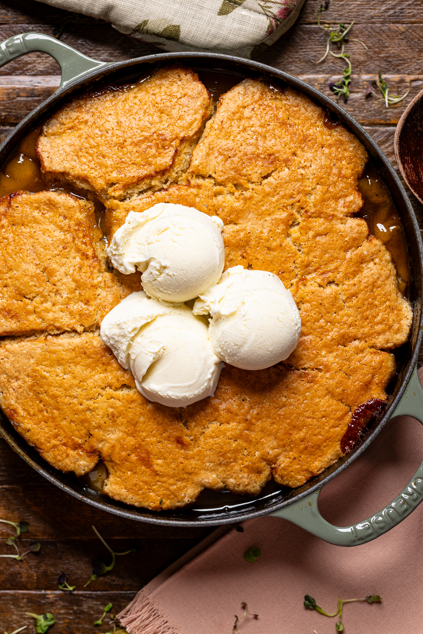 Image of peach cobbler in a skillet on a brown wood table with scoops of ice cream.