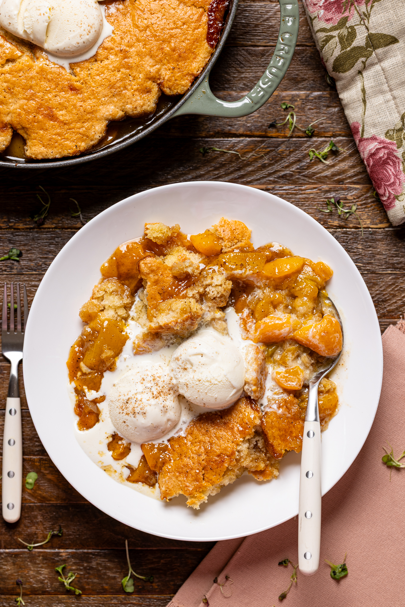 Image of peach cobbler in low bowl with scoops of ice cream with a spoon.