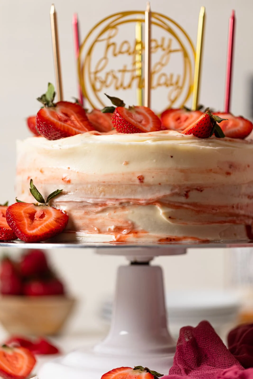 Strawberry shortcake birthday cake with sliced strawberries on top, birthday candles, on a cake stand on a white table.