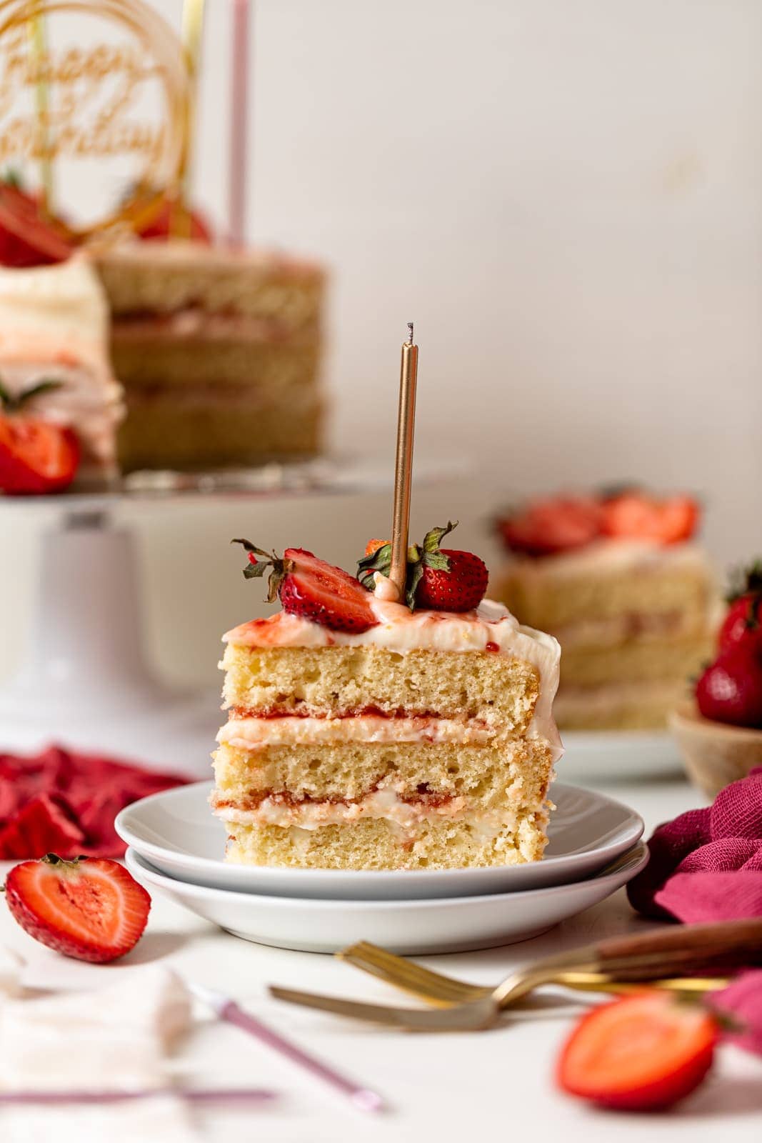 Slice of cake on stacked white plates with sliced strawberries and the full cake on a cake stand in the background; all on a white table. 
