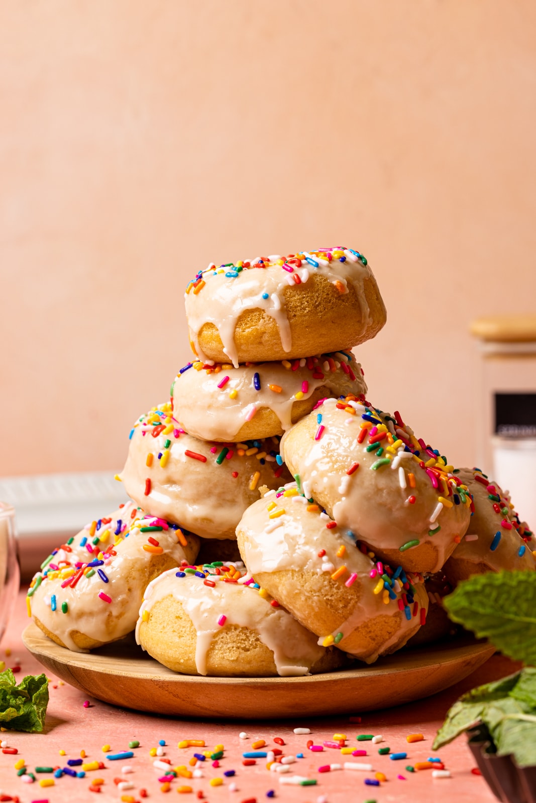 Stack of donuts on a plate on a pink table with sprinkles and garnish of herbs.