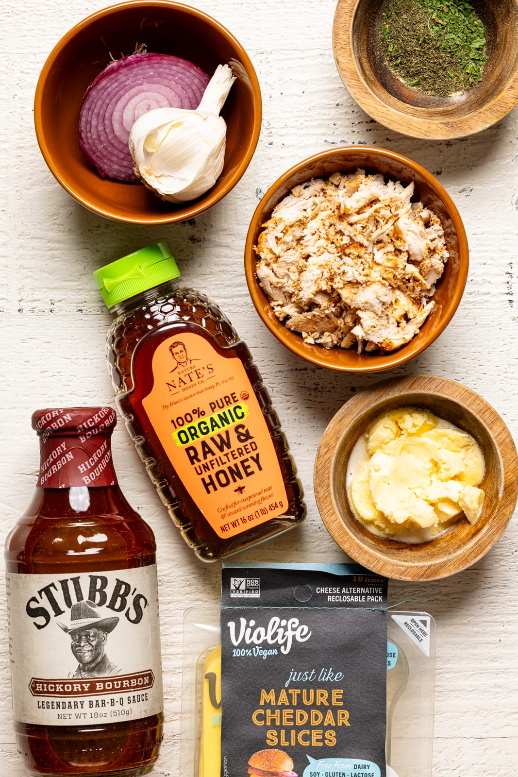 Ingredients on a white table including bbq sauce, honey, cheese slices, chicken, butter, onions, garlic, and herbs + seasonings. 