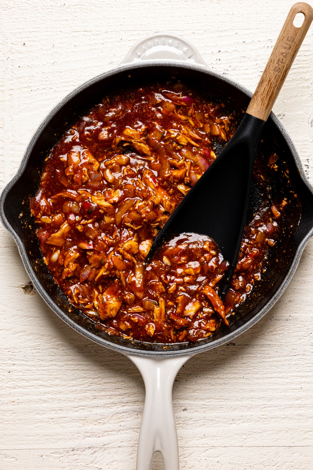 Shredded bbq chicken in a white skillet on a white table with a black spoon.