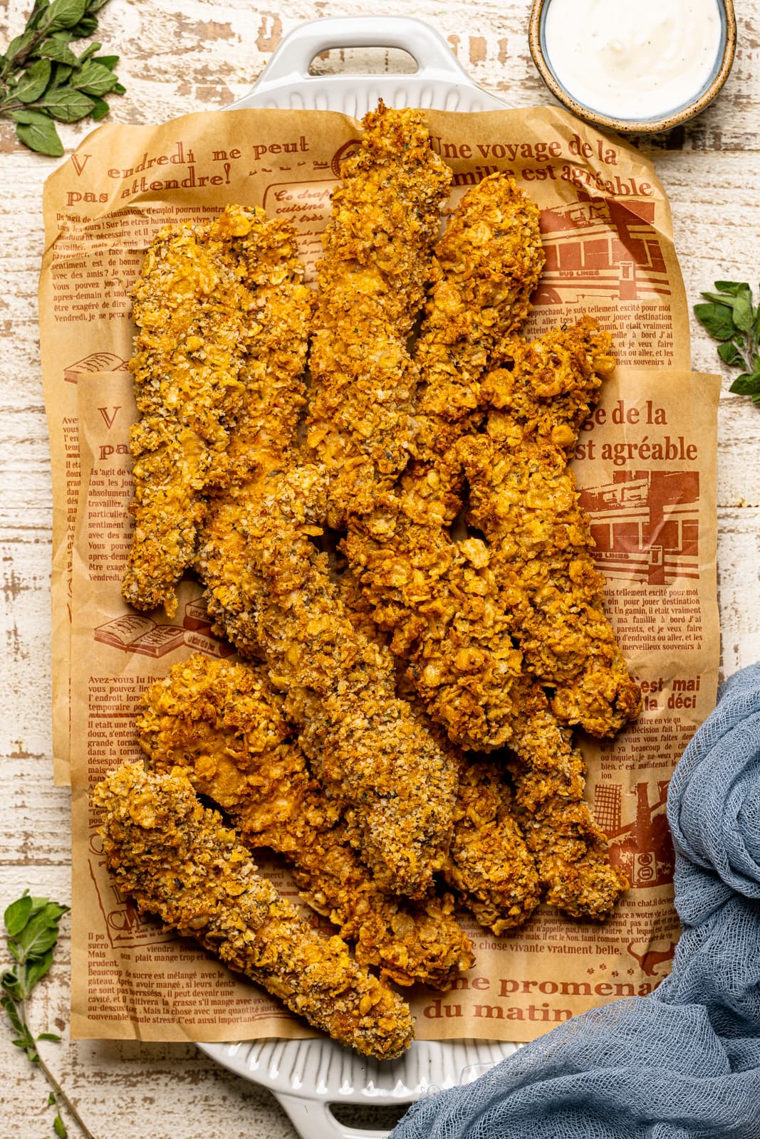 Crispy chicken tenders piled on a platter with brown sandwich paper on a white wood table and a blue napkin.