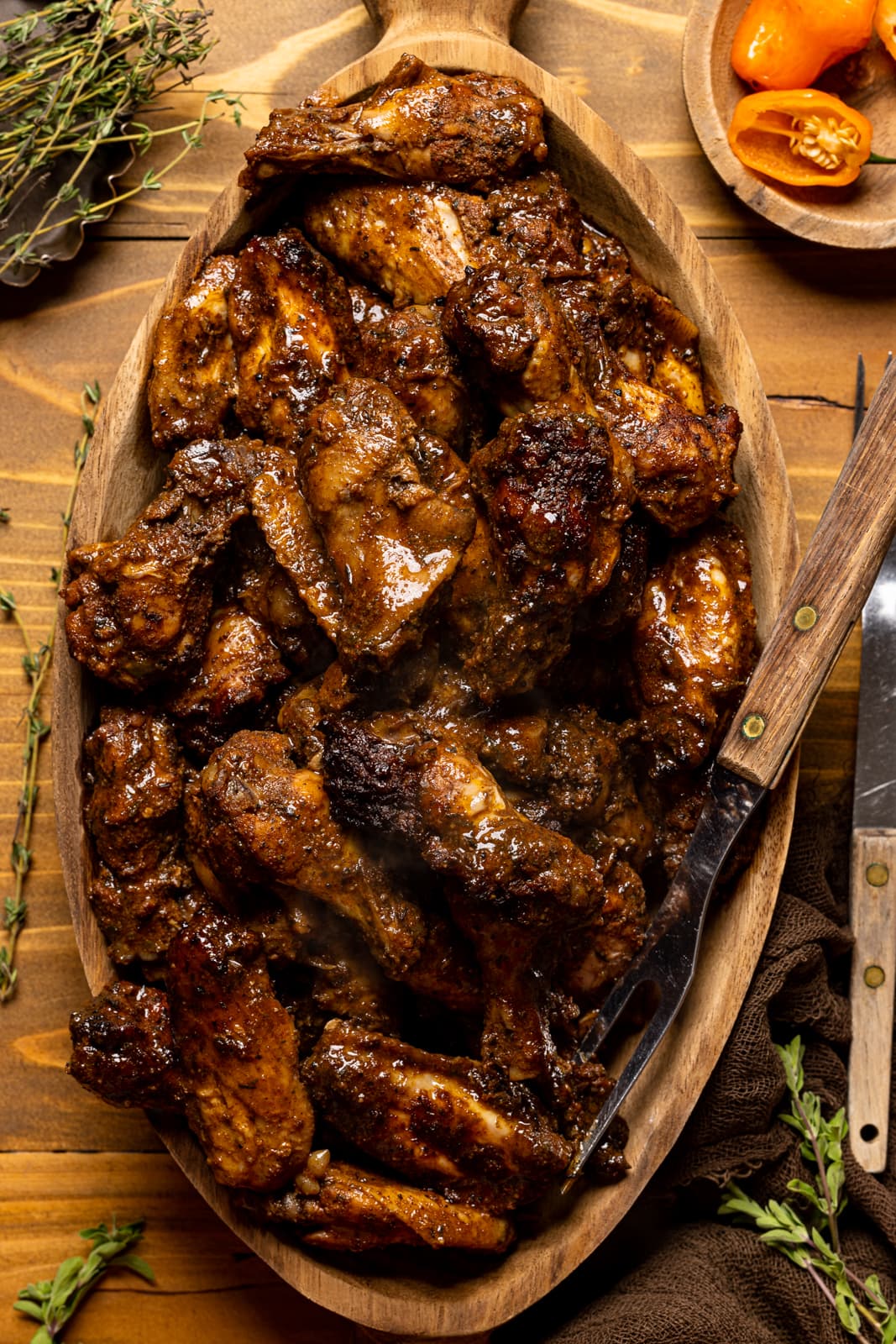 Chicken wings in a brown wood serving bowl with two forks, thyme sprigs, and peppers on a brown wood table.