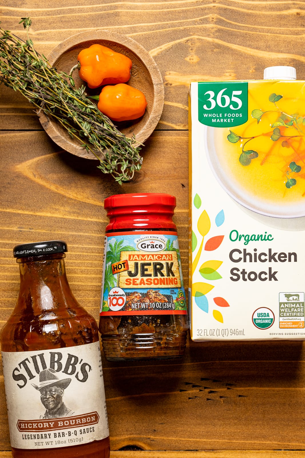 Ingredients on a brown wood table including jerk seasoning, bbq sauce, chicken stock, thyme sprigs, and peppers.