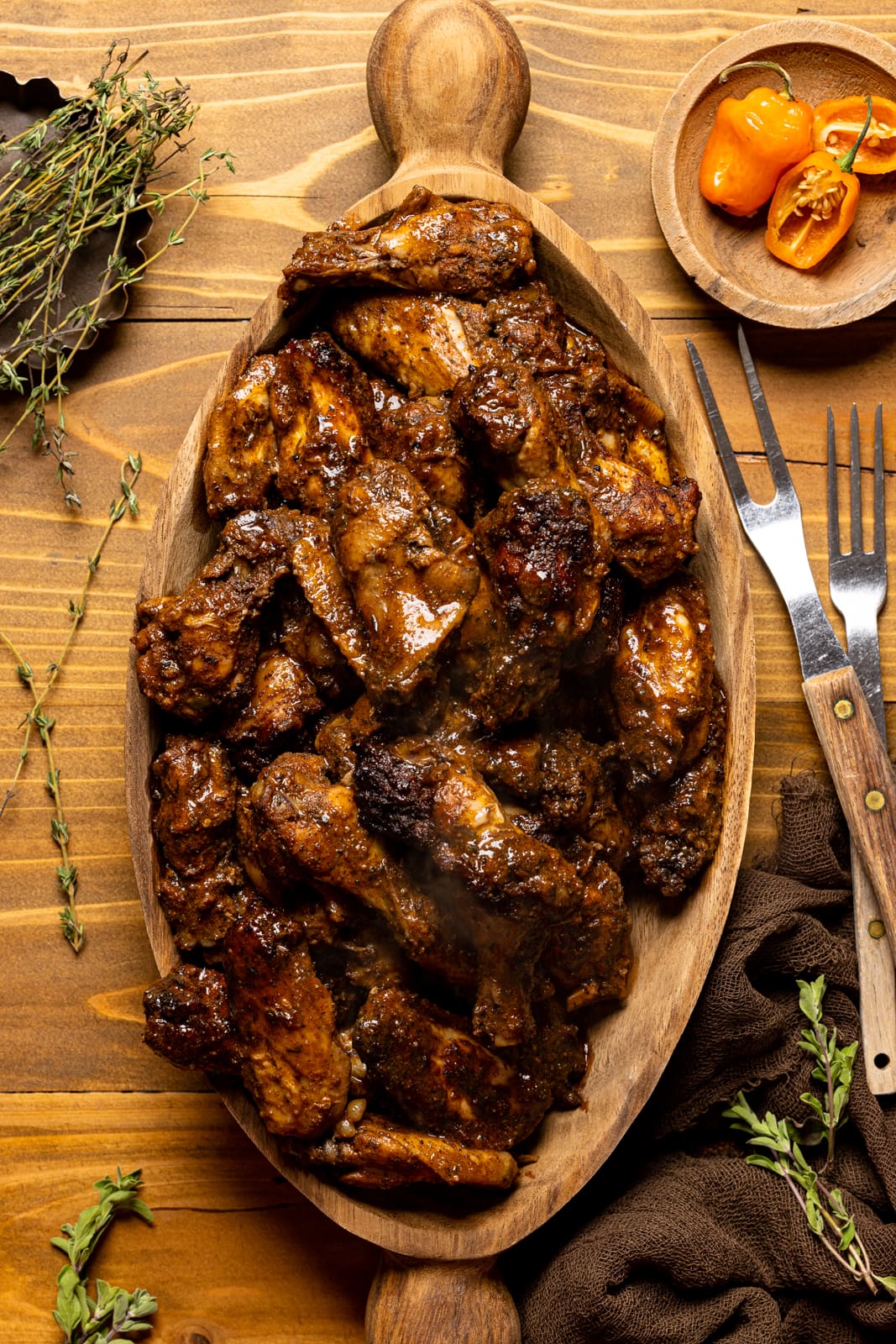 Chicken wings in a brown wood serving bowl with two forks, thyme sprigs, and peppers on a brown wood table.