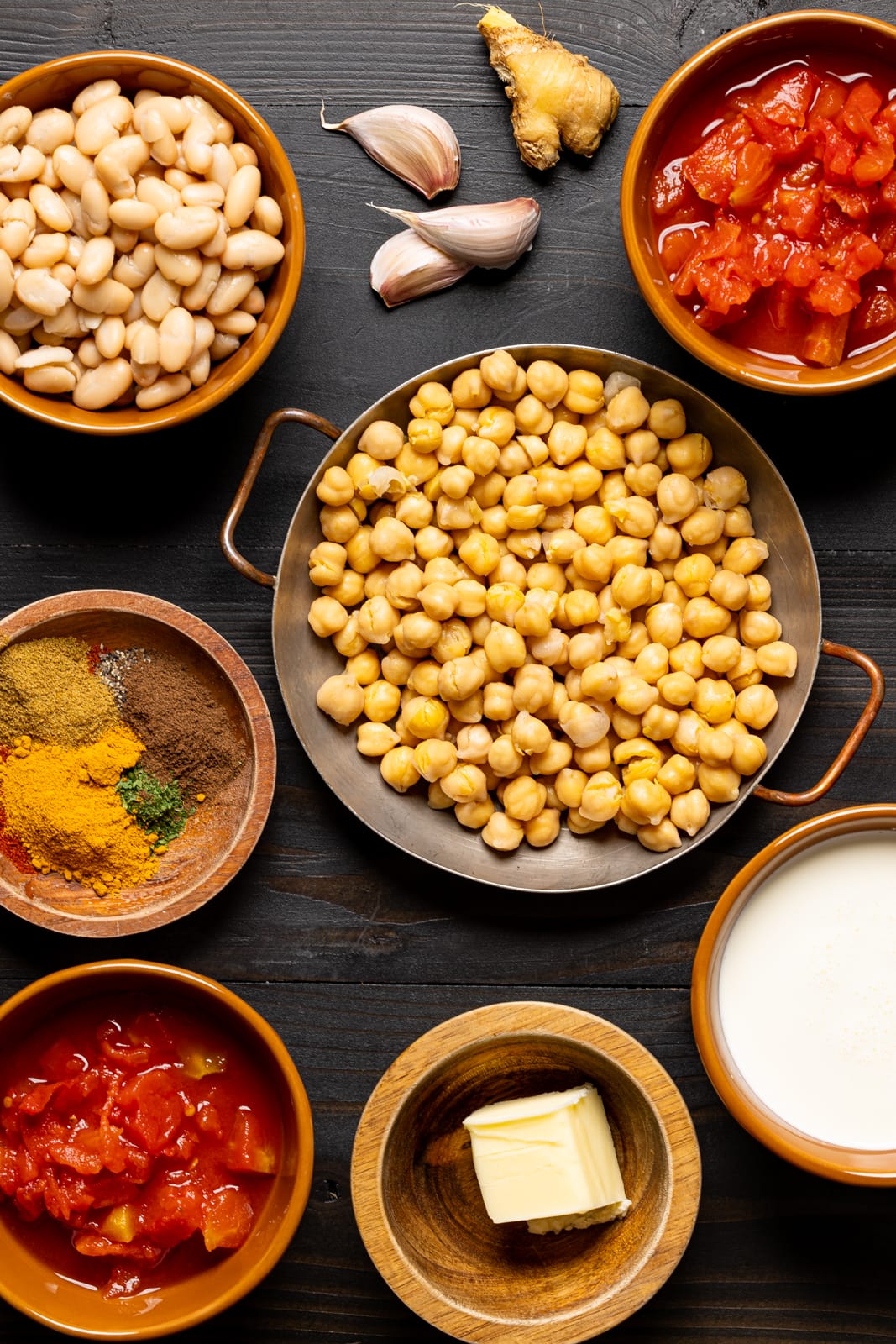 Ingredients on a black wood table including chickpeas, white beans, herbs and spices, tomatoes, butter, garlic, ginger, and heavy cream.