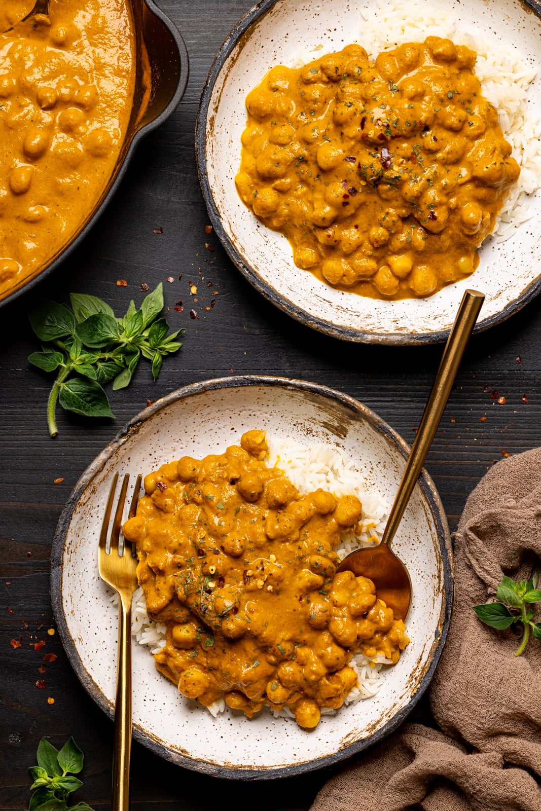 Two low bowls of chickpeas on a black wood table with a brown napkin and herbs and a fork and spoon.