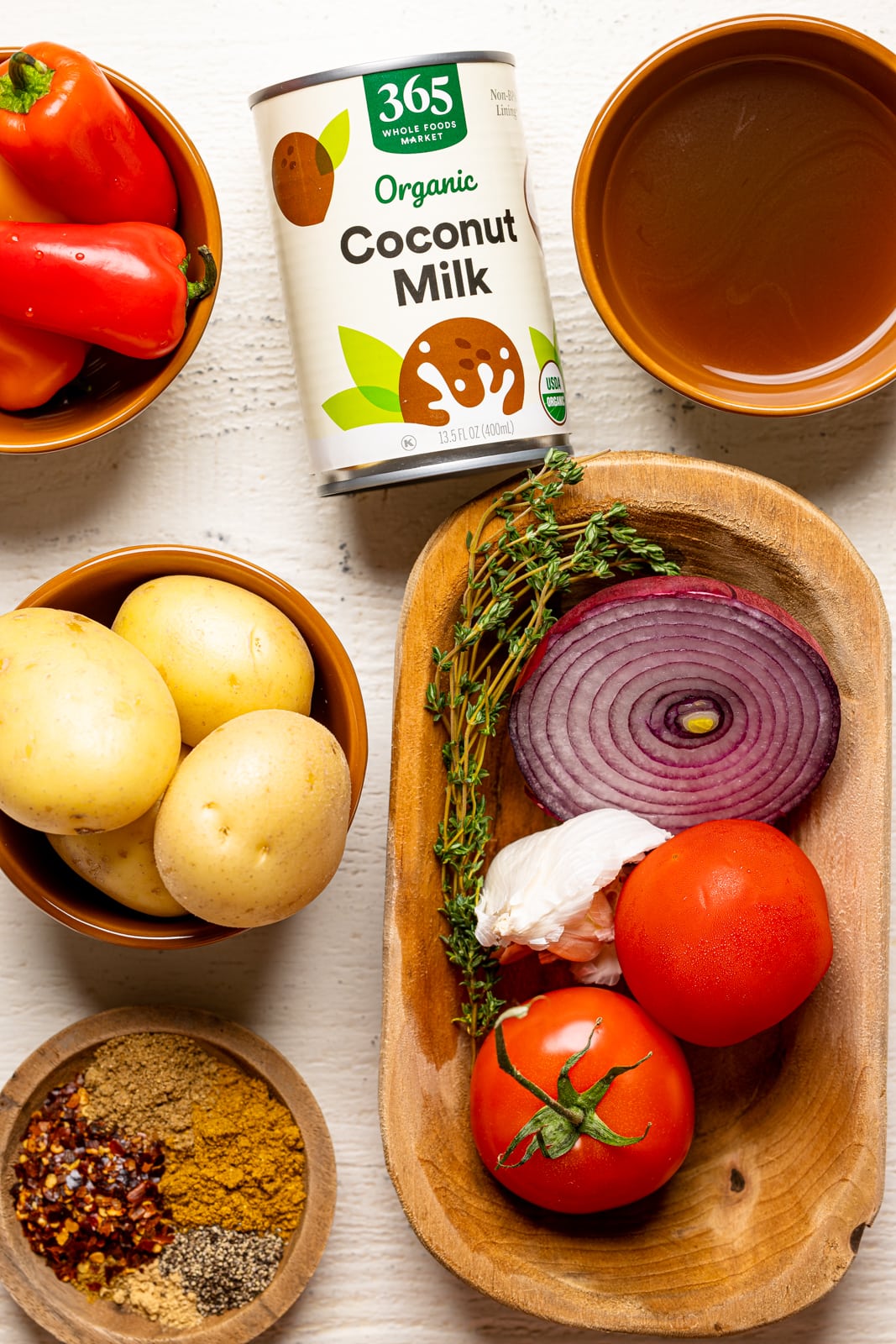 Ingredients on a white table including peppers, tomatoes, onions, herbs + seasonings, thyme sprigs, potatoes, coconut milk, and chicken stock.