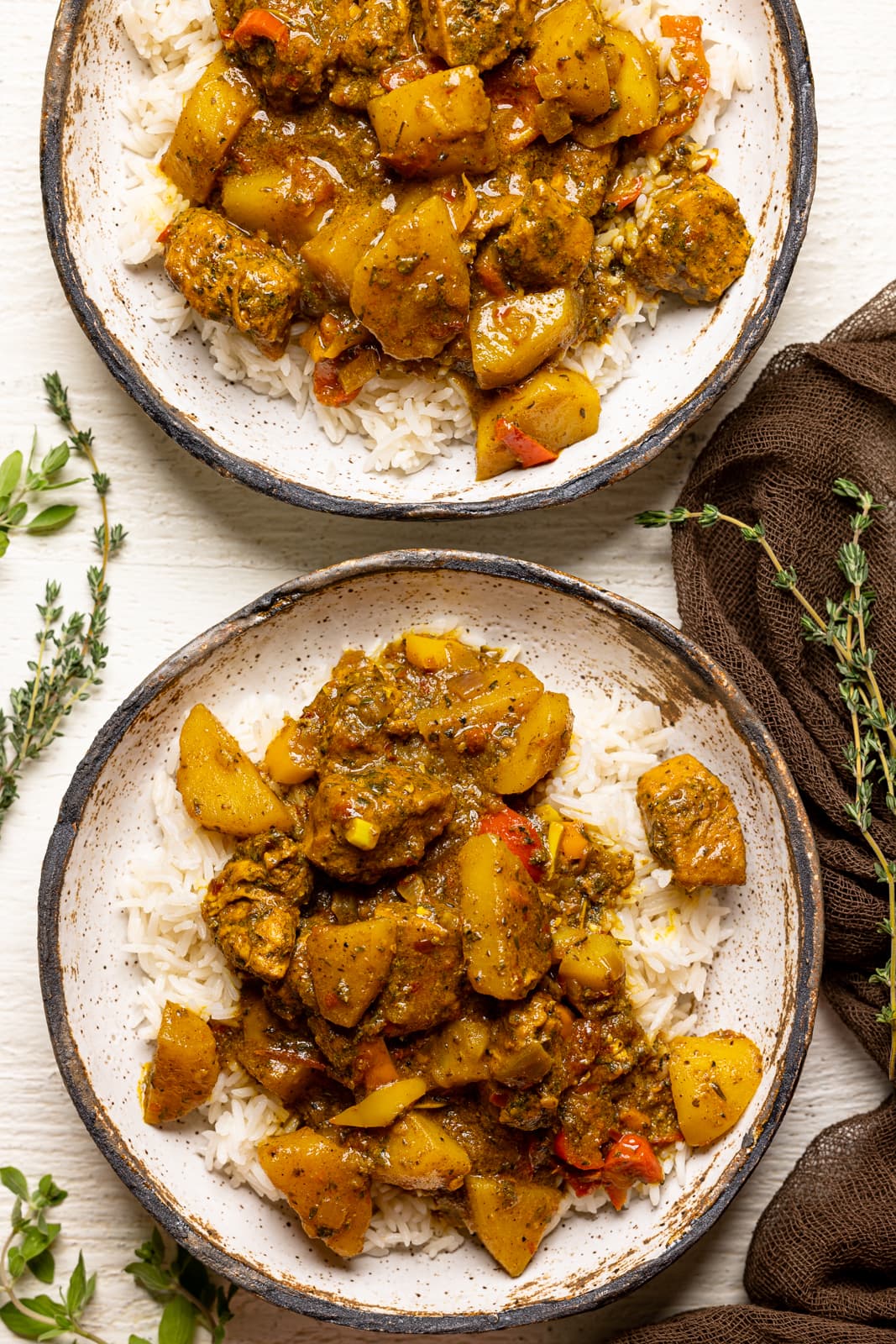 Two bowls of curry chicken with white rice on a white table with a brown napkin and herbs.