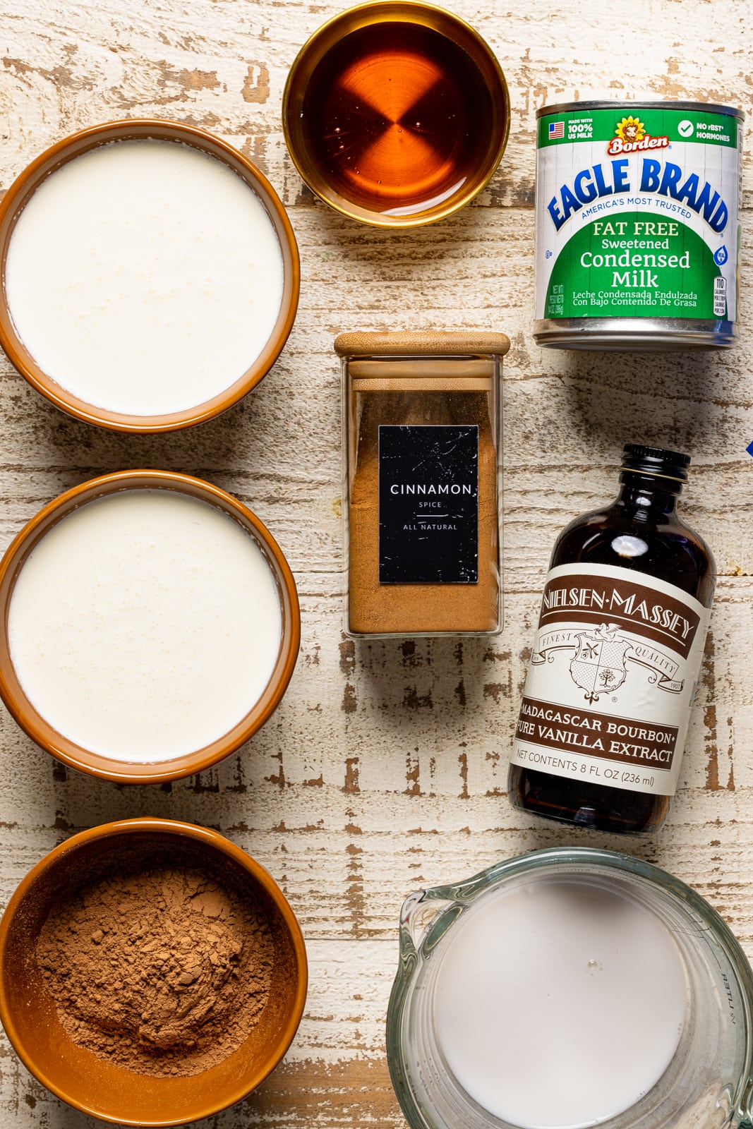 Ingredients on a white wood table including condensed milk, cocoa powder, milk, heavy cream, vanilla, cinnamon, and maple syrup.