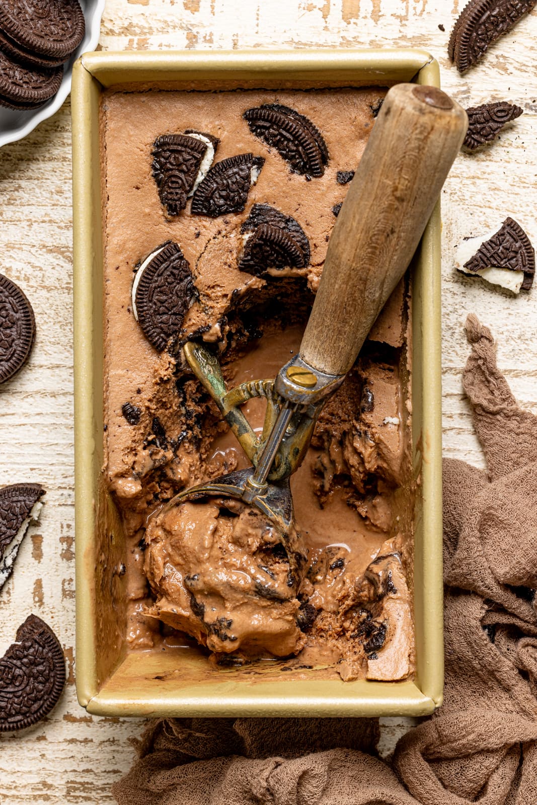 Chocolate Oreo ice cream in a gold tin on a white wood table with a brown napkin and oreo cookies.