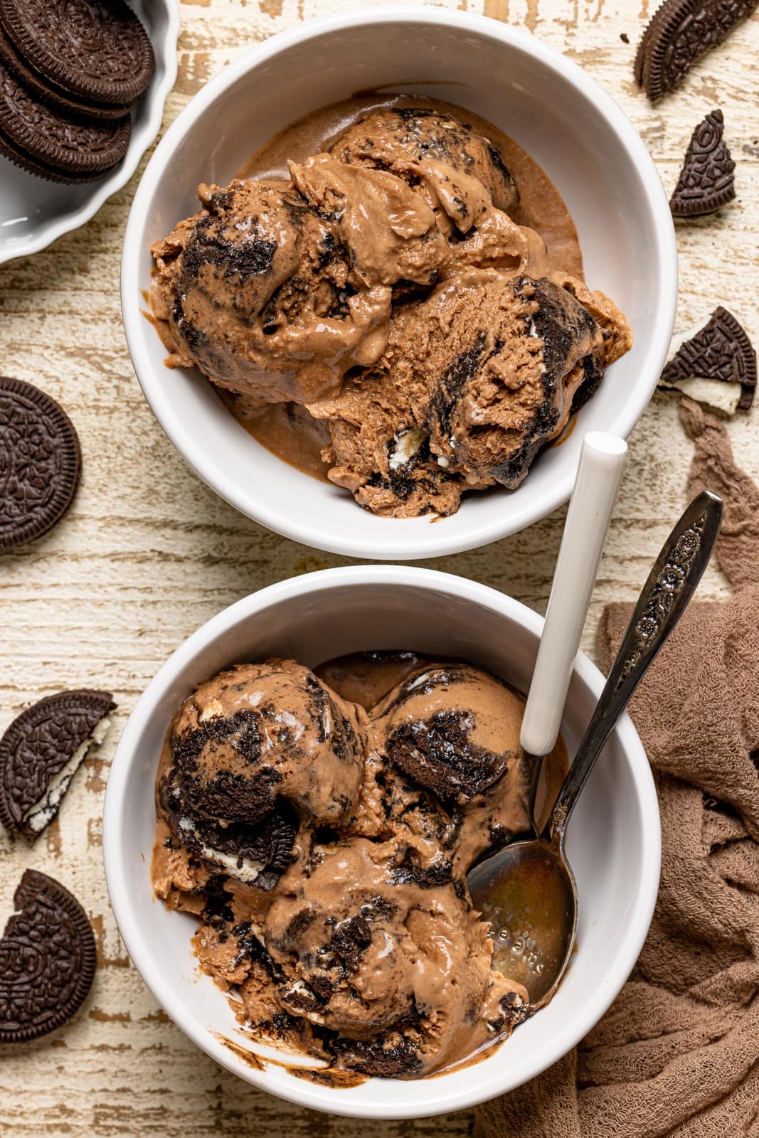 Two white bowls of ice cream on a white wood table with a brown napkin and oreo cookies.