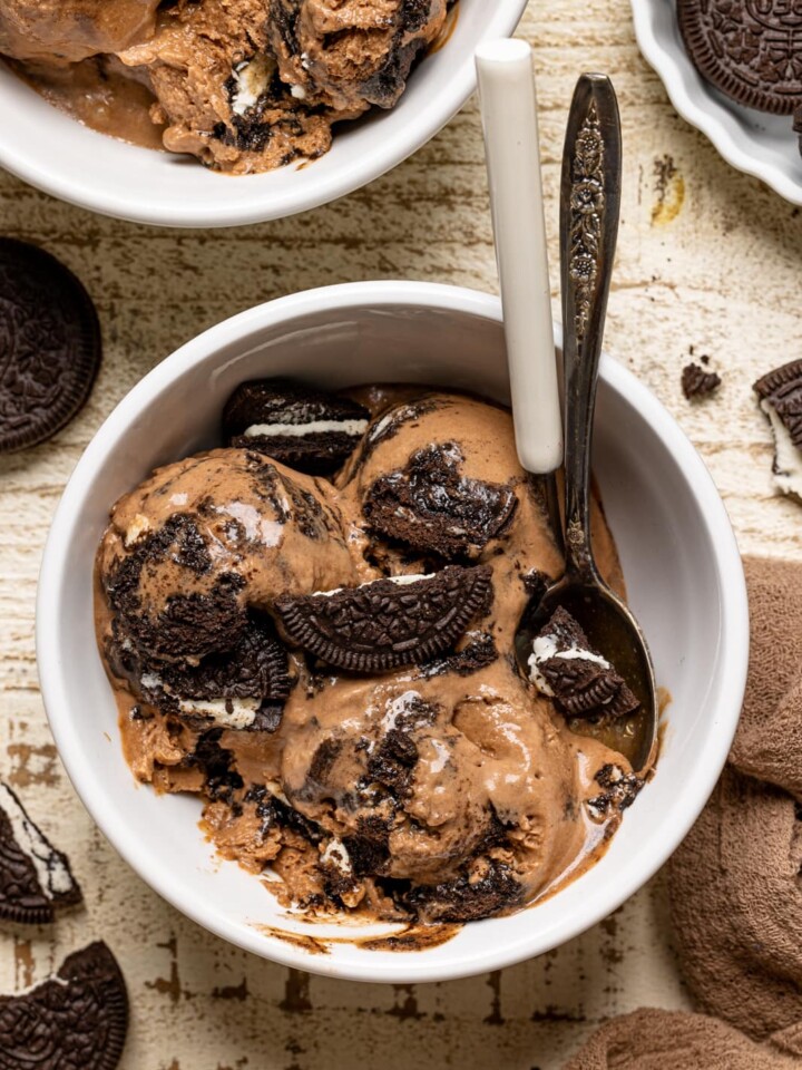 Chocolate ice cream in a white bowl on a white wood table with two spoons and Oreo cookies.