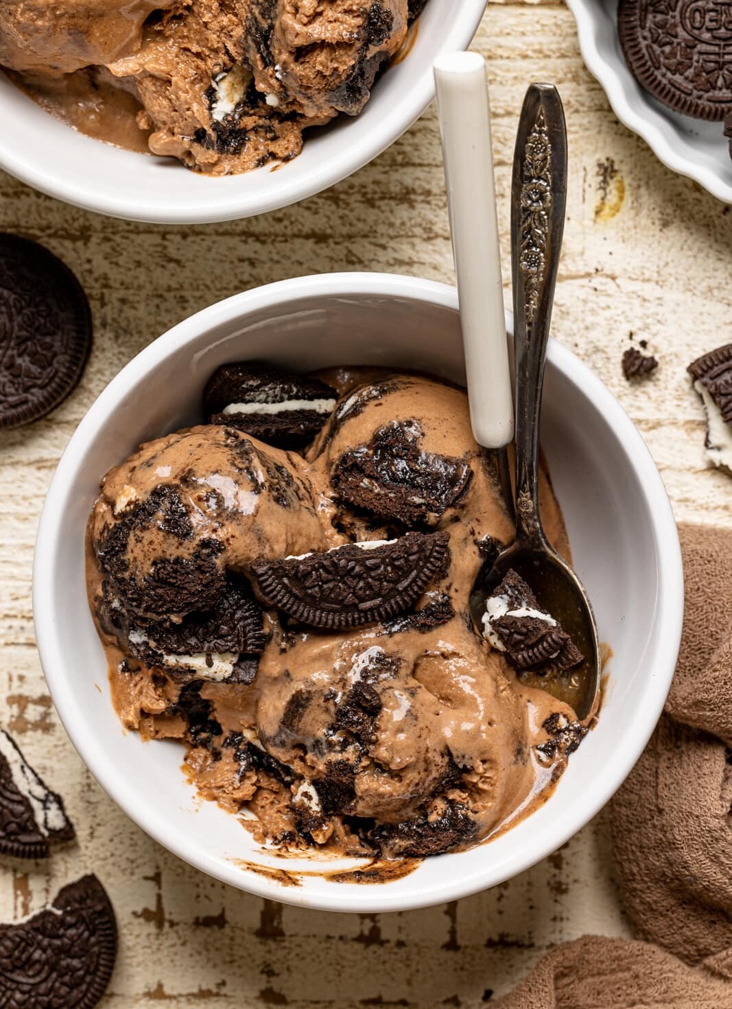 Chocolate ice cream in a white bowl on a white wood table with two spoons and Oreo cookies.