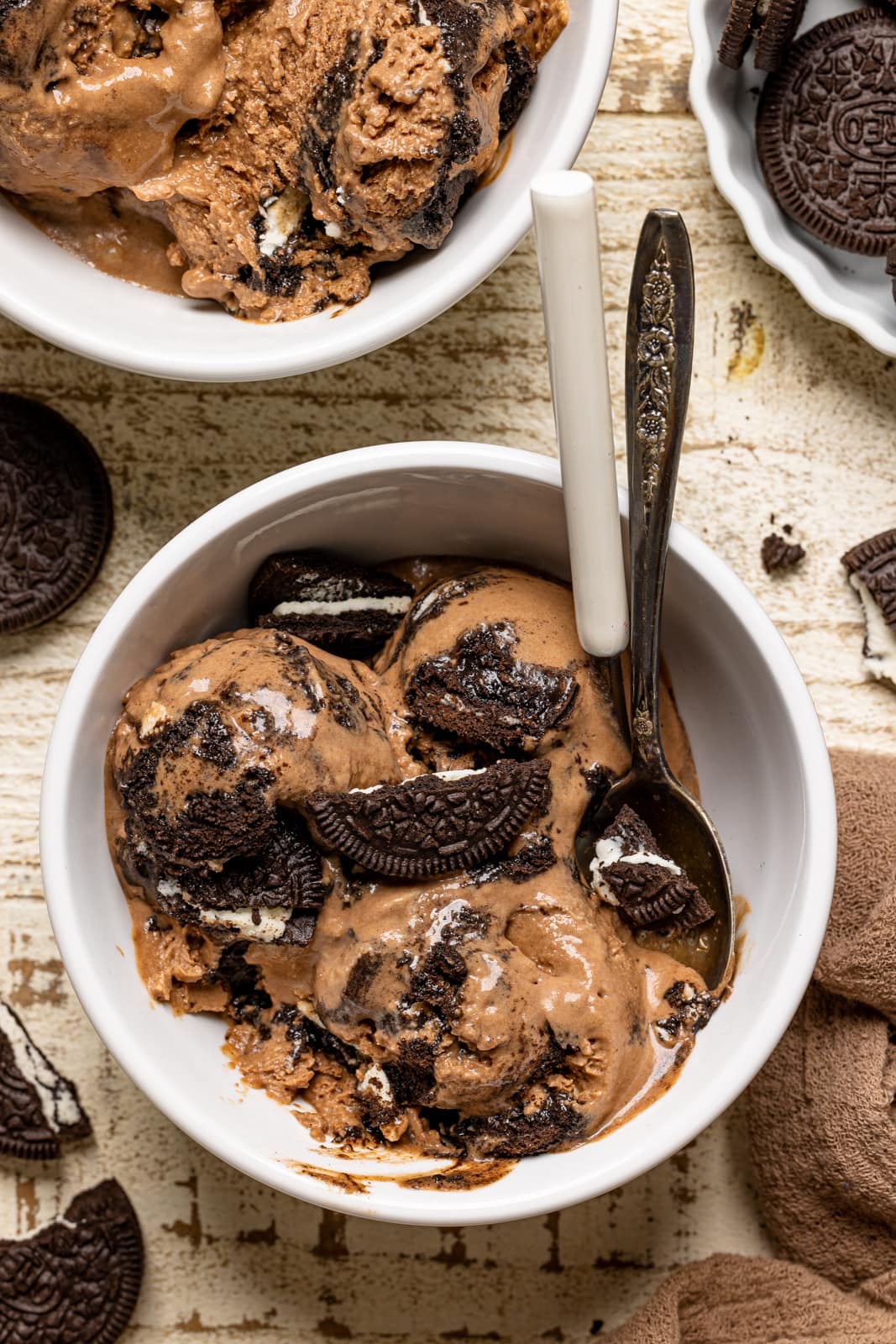 Chocolate ice cream in a white bowl on a white wood table with two spoons and Oreo cookies.