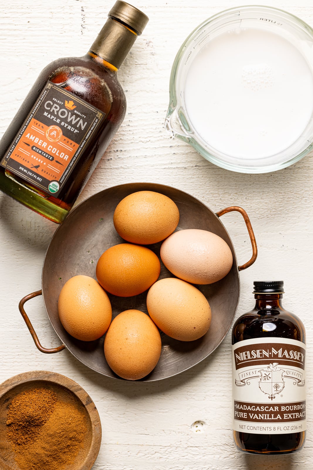 Ingredients on a white table including maple syrup, eggs, milk, vanilla, and cinnamon.