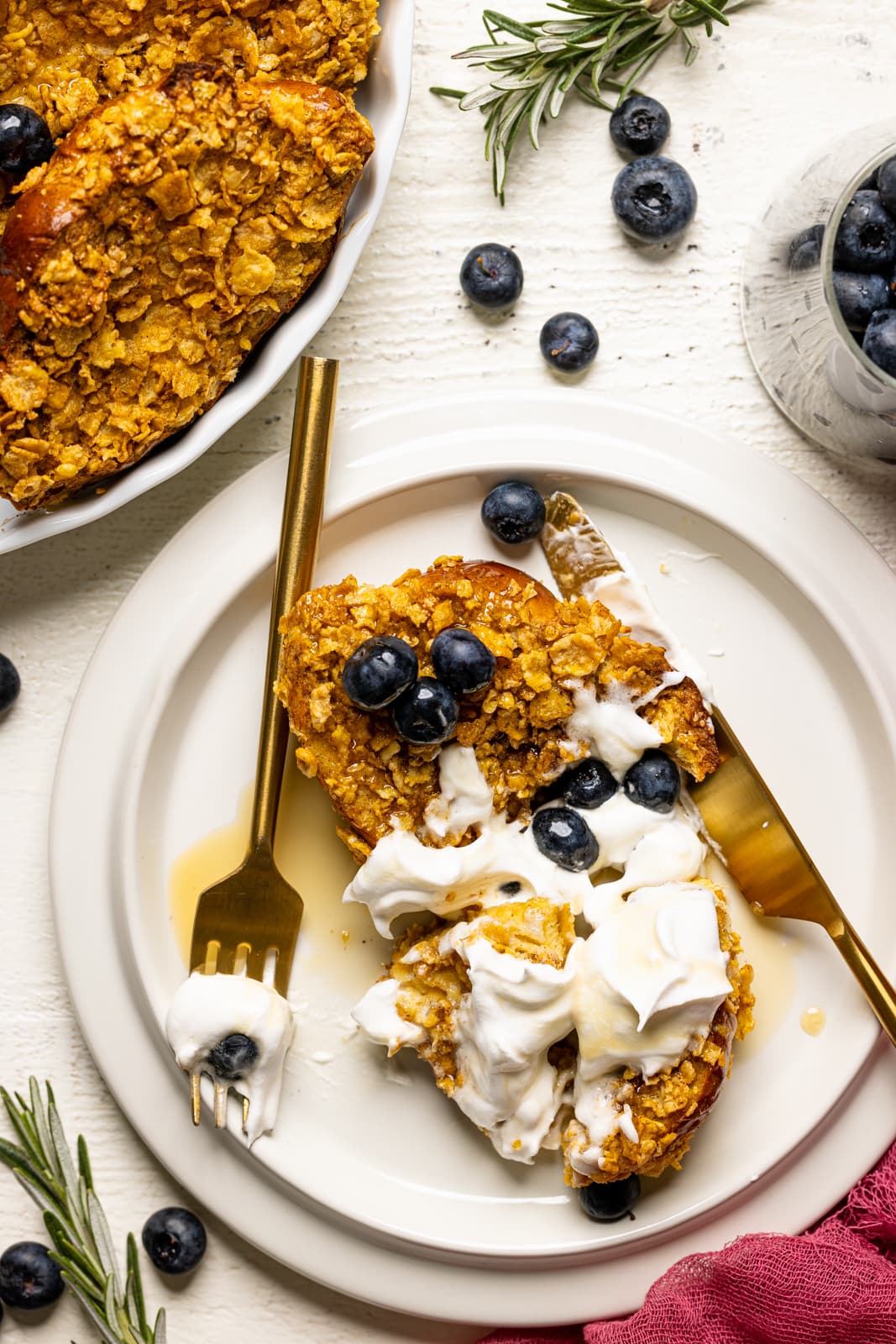 Slice of french toast on a white plate with blueberries, whipped cream, fork + knife, on a white table.