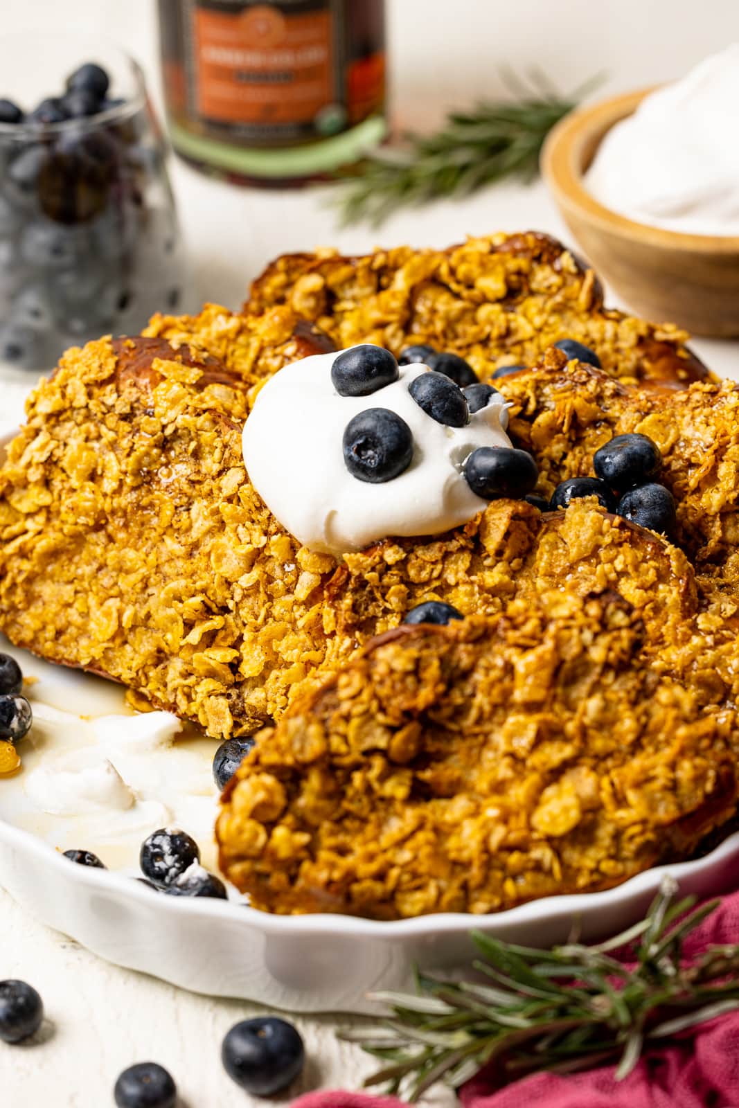 French toast in a white baking dish on a white table with blueberries, whipped cream, and herbs.