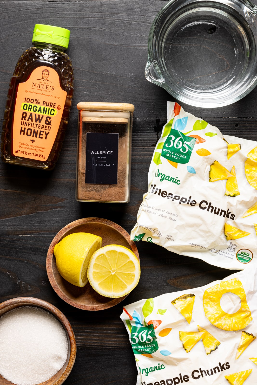 Ingredients on a black table including frozen pineapples, honey, lemons, water, cinnamon, and cane sugar.