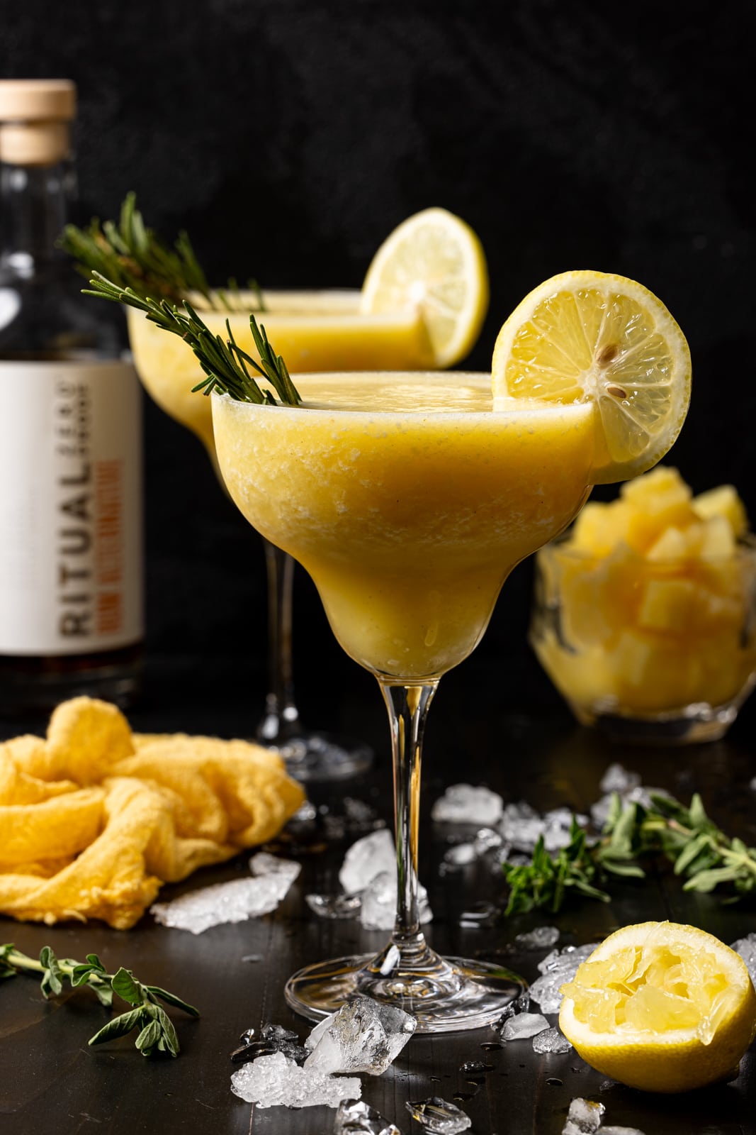 Two glasses of frozen margarita with a yellow napkin, lemon slices, pineapples, and alcohol alternative. 