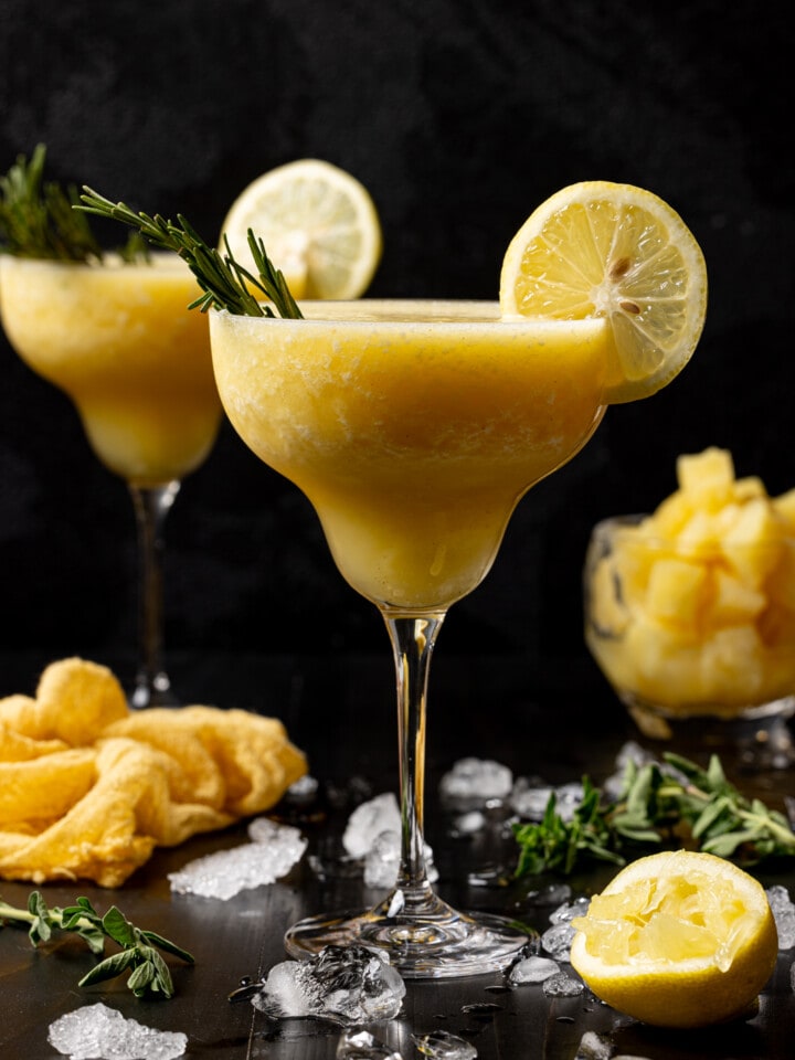 Two glasses of frozen margaritas with ice, lemon slices, and pineapple chunks.