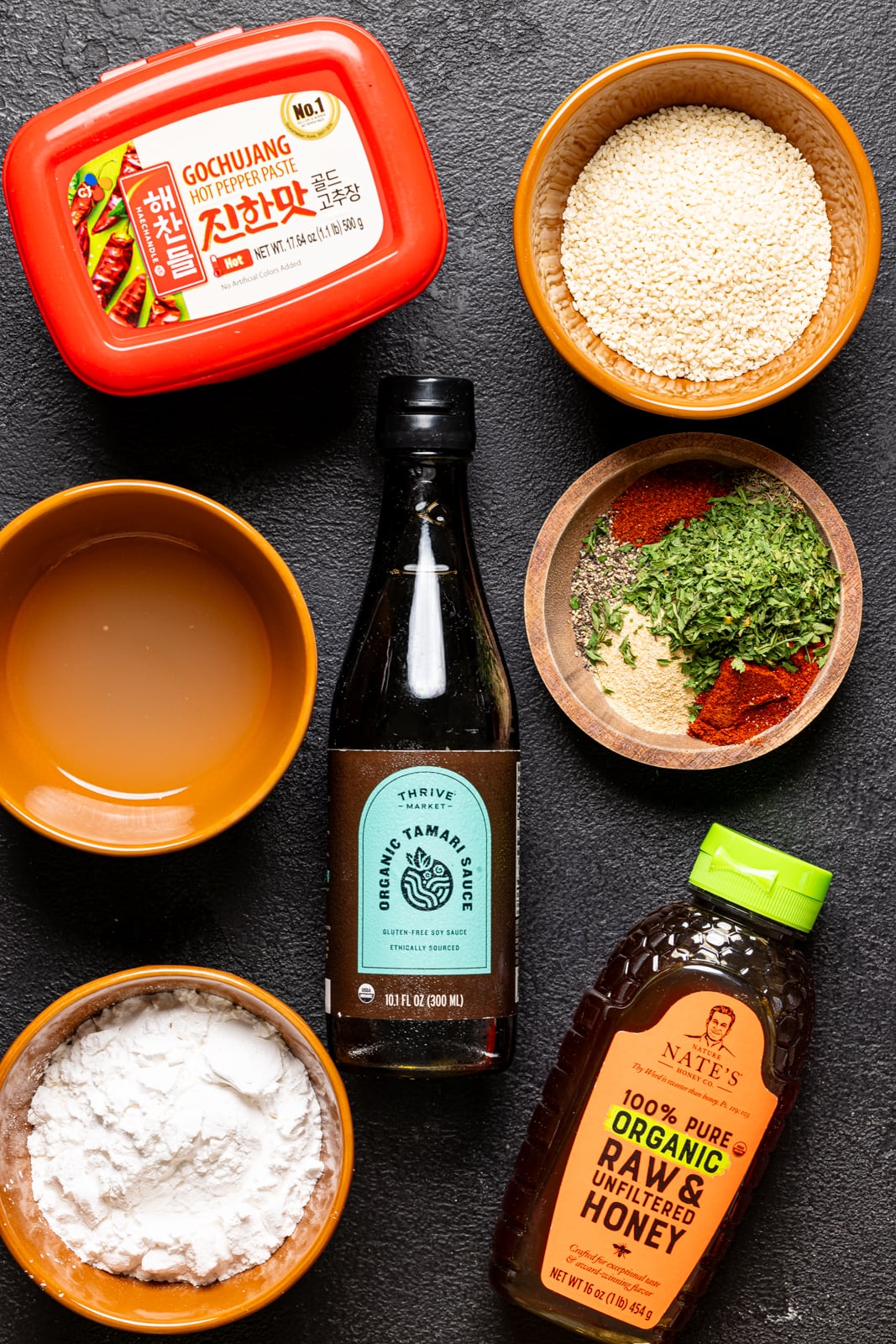 Ingredients on a black table including gochujang paste, apple cider vinegar, cornstarch, honey, tamarind sauce, sesame seeds, and herbs + seasonings. 