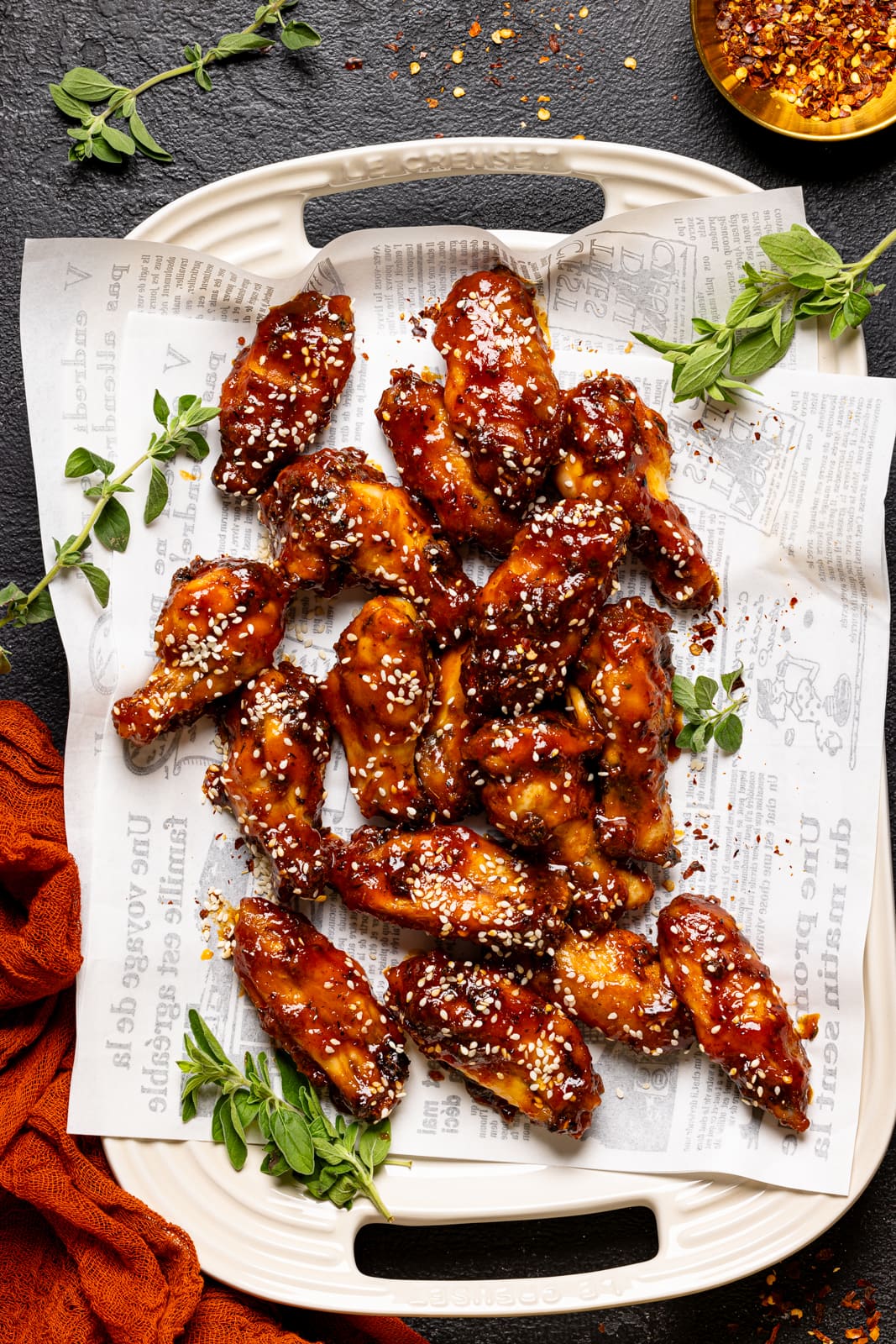 Chicken wings on a platter with white paper, a red napkin, red pepper flakes, and herbs.