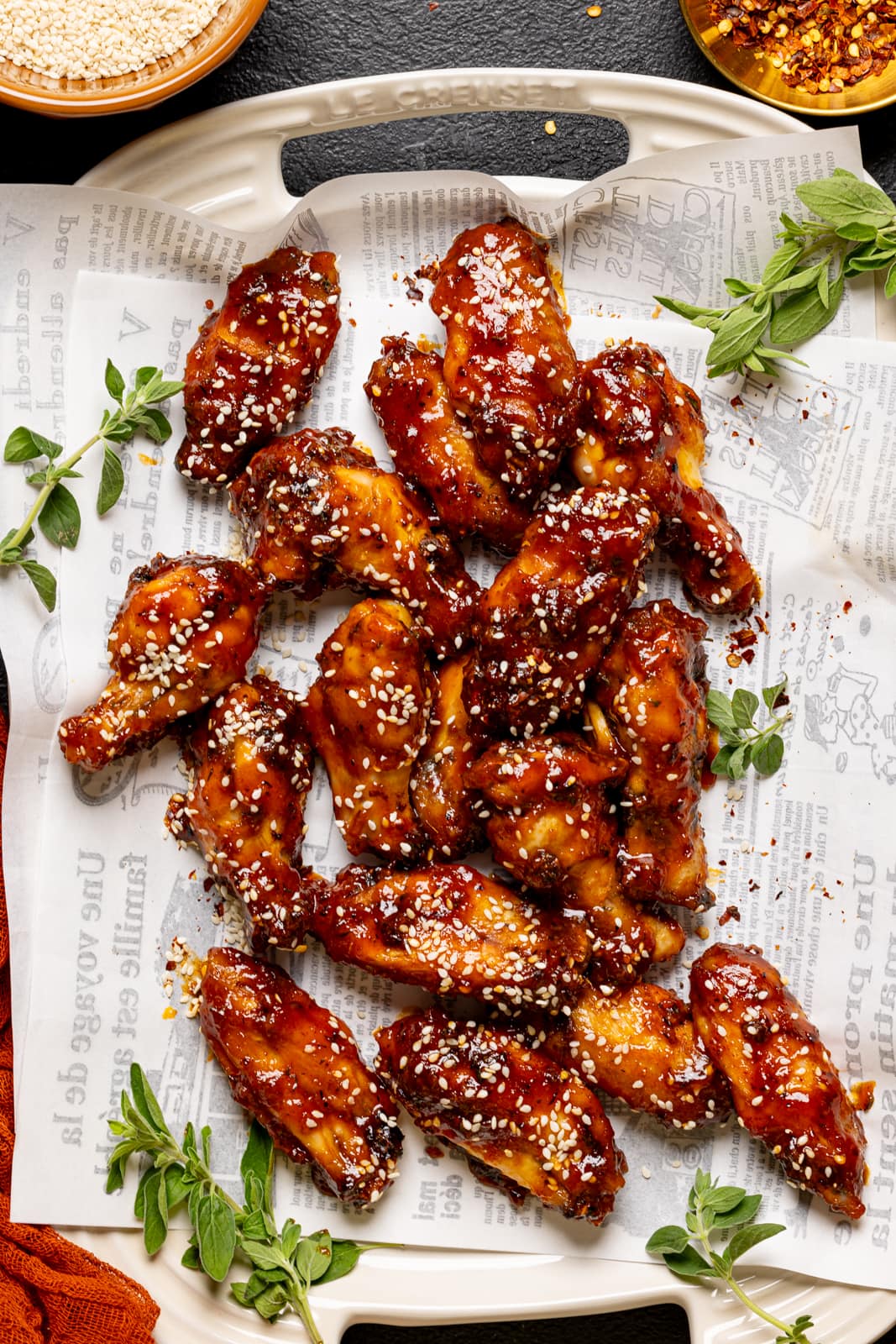 Up close of chicken wings on a platter with white paper, herbs, + sesame seeds on a black table.