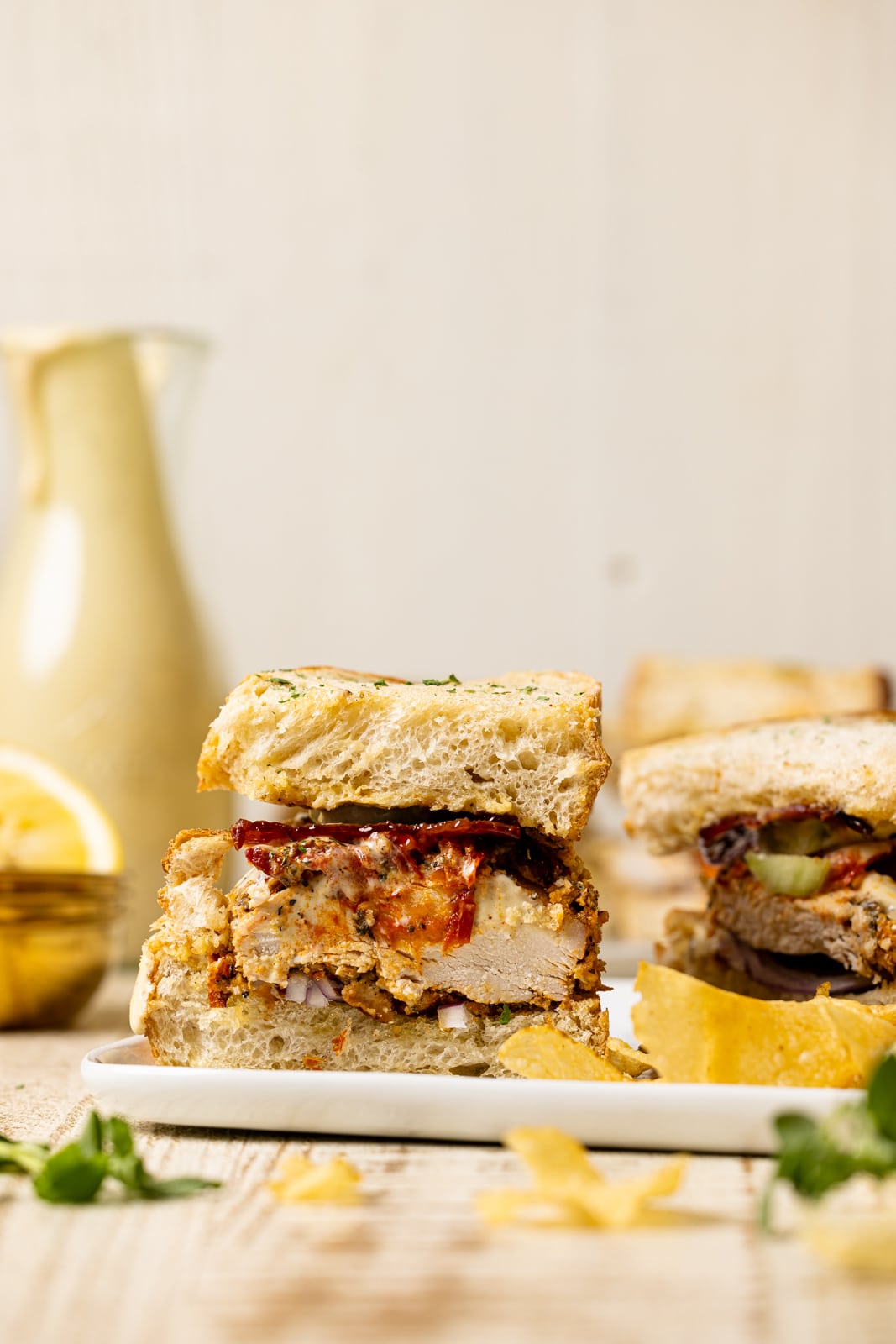 Two sliced sandwiches on a white plate with chips and honey mustard dressing in the background. 