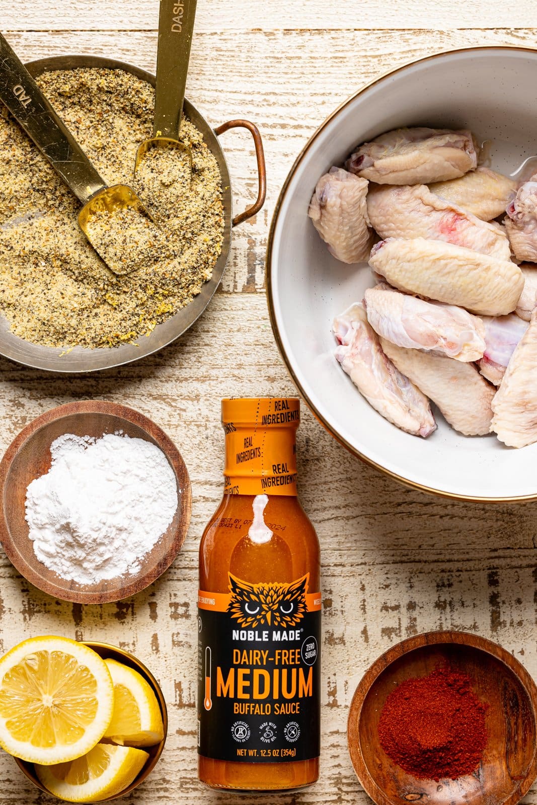 Ingredients on a white wood table including chicken wings, buffalo sauce, lemon pepper seasoning, lemons, cornstarch, and seasoning.