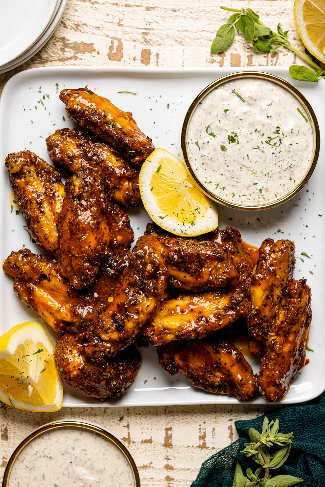 Chicken wings on a white plate with dipping sauces and lemon slices on a white table with herbs.