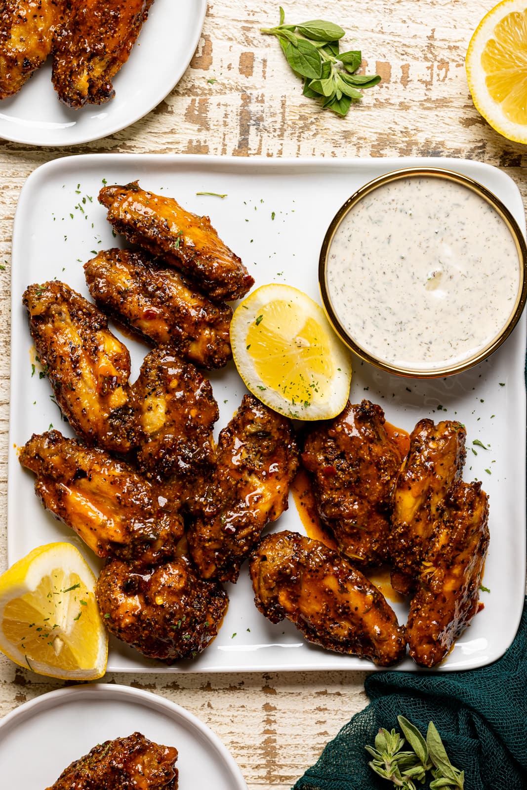 Chicken wings on a white plater with lemon slices, dip, and plates of wings on a white wood table.