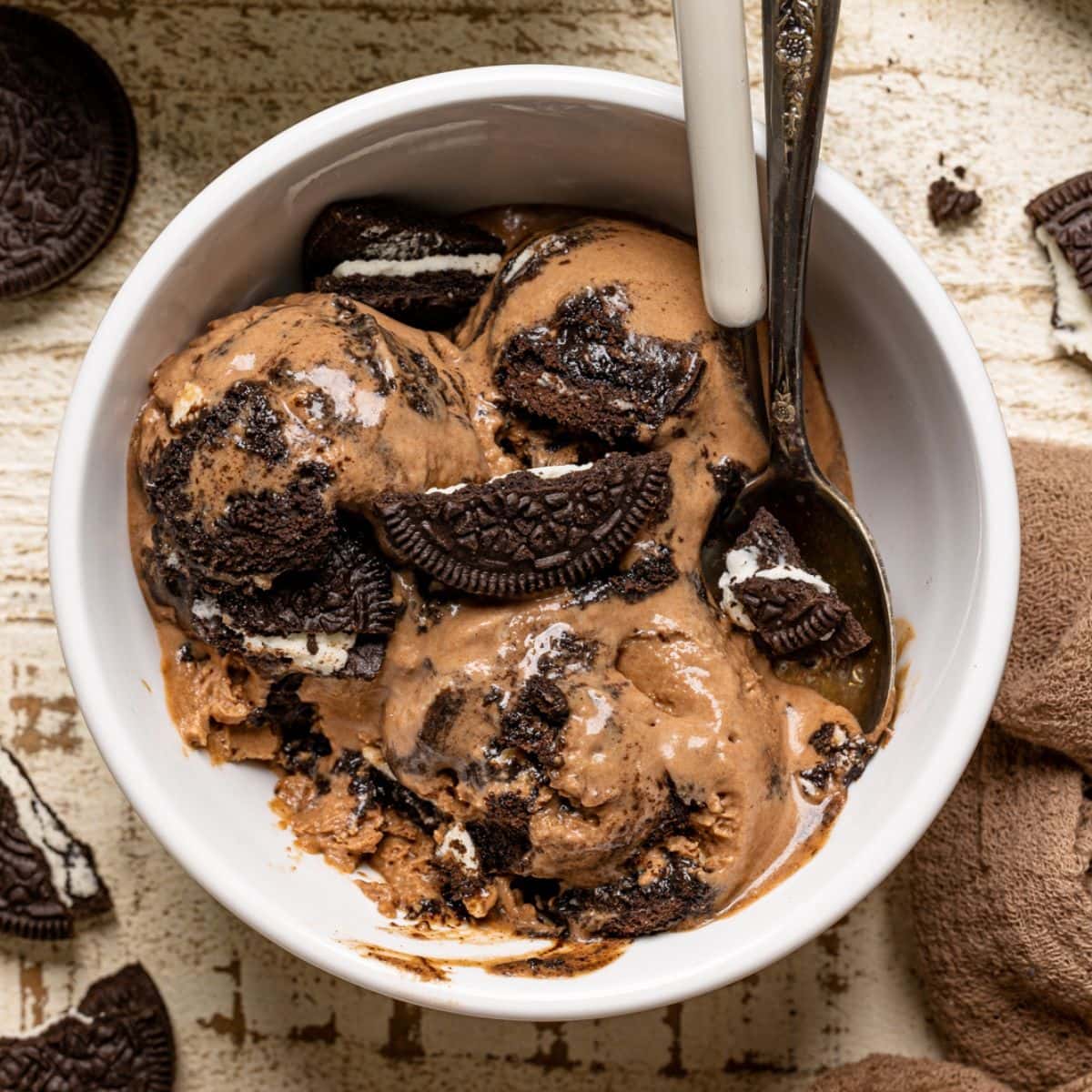 Ice cream in a white bowl on a white wood table with two spoons and oreo cookies.