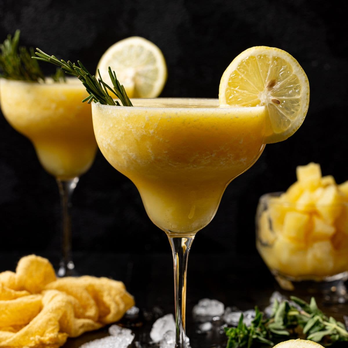 Two glasses of frozen margaritas with ice, lemon slices, and pineapple chunks.