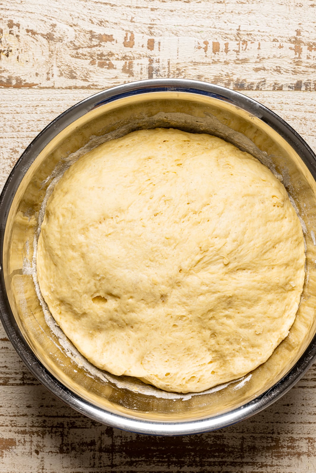 Donut dough risen in a large silver bowl on a white wood table.