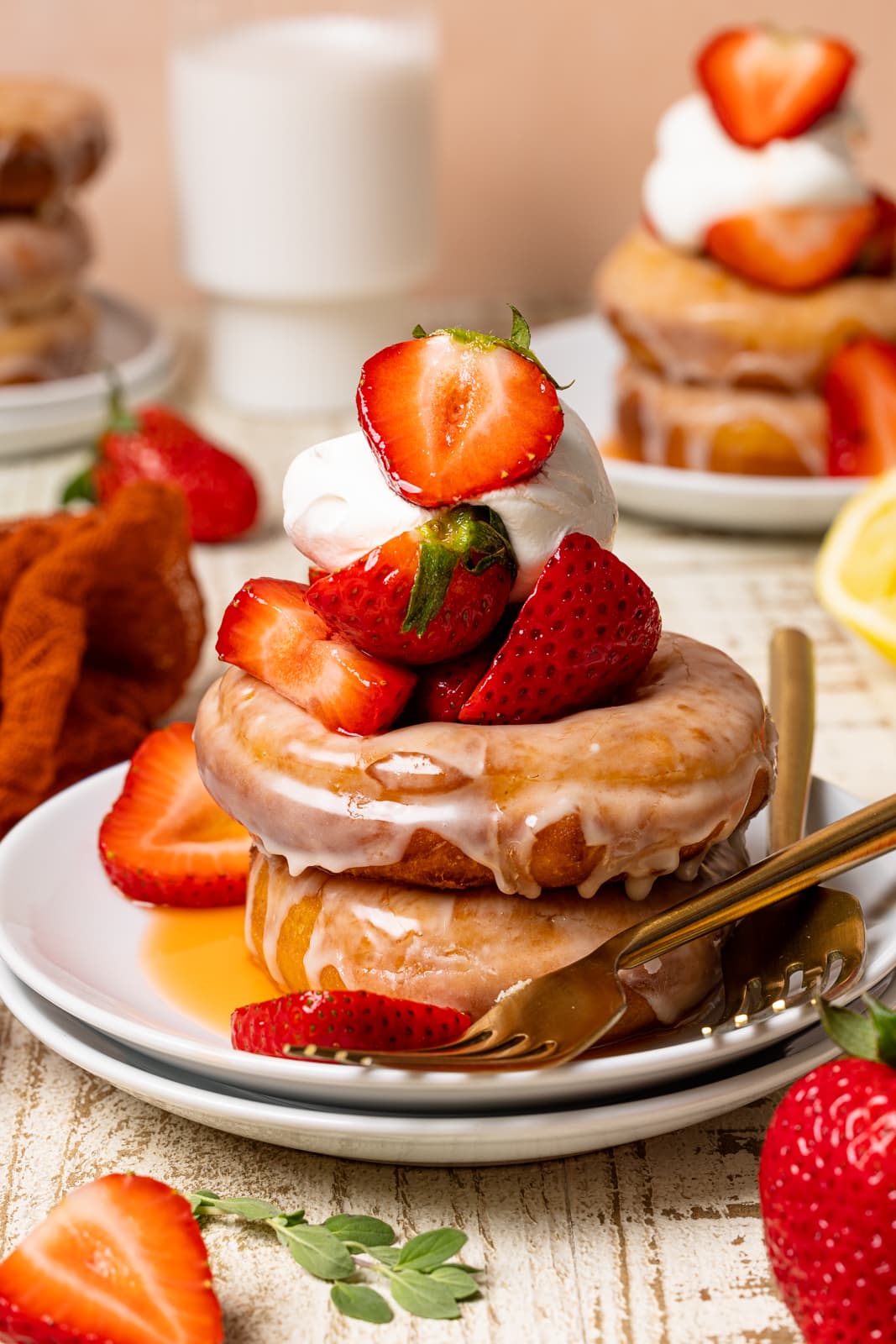 Stacked donuts on two white plates with strawberries, whipped cream, two forks, and milk in the background on a white wood table.