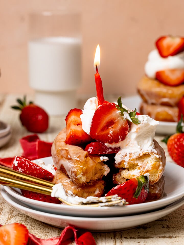 Two donuts stacked on two plates with strawberries, whipped cream, and a birthday candle on a white wood table with a glass of milk in the background and strawberries.