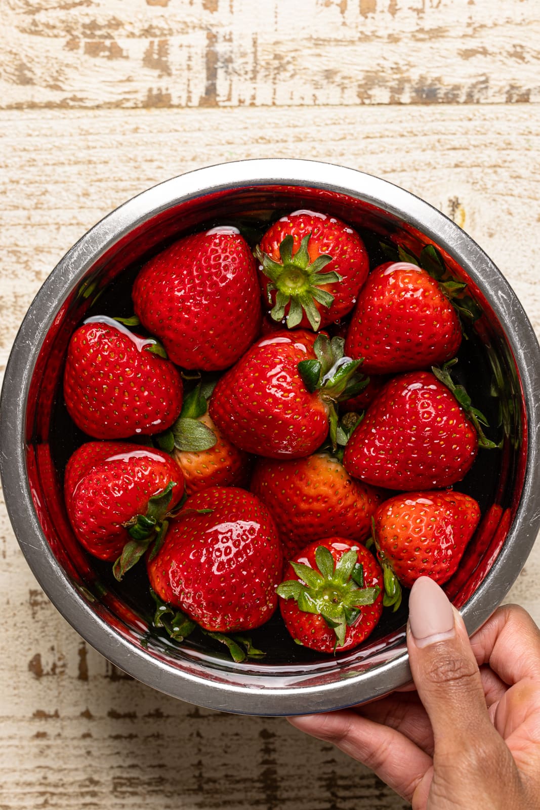 Strawberries soaked in water and vinegar in a silver bowl on a white wood table.