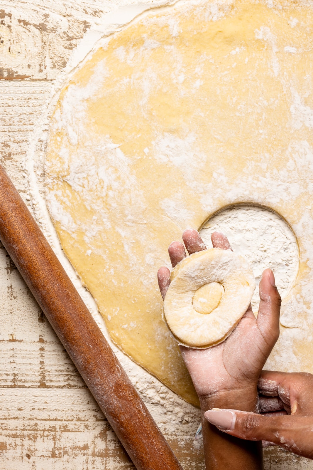 One donut cut out of dough unto a white wood table with a brown wood roller.